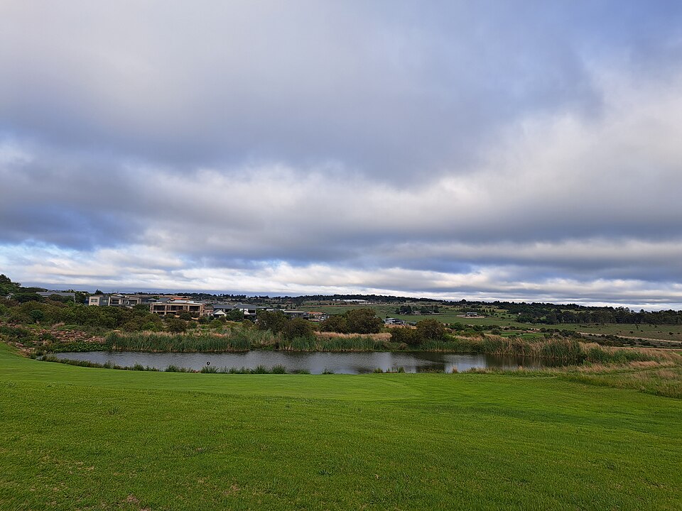 A fairway in the foreground with a reed-fringed dam beyond it and houses on the horizon under a cloudy sky.