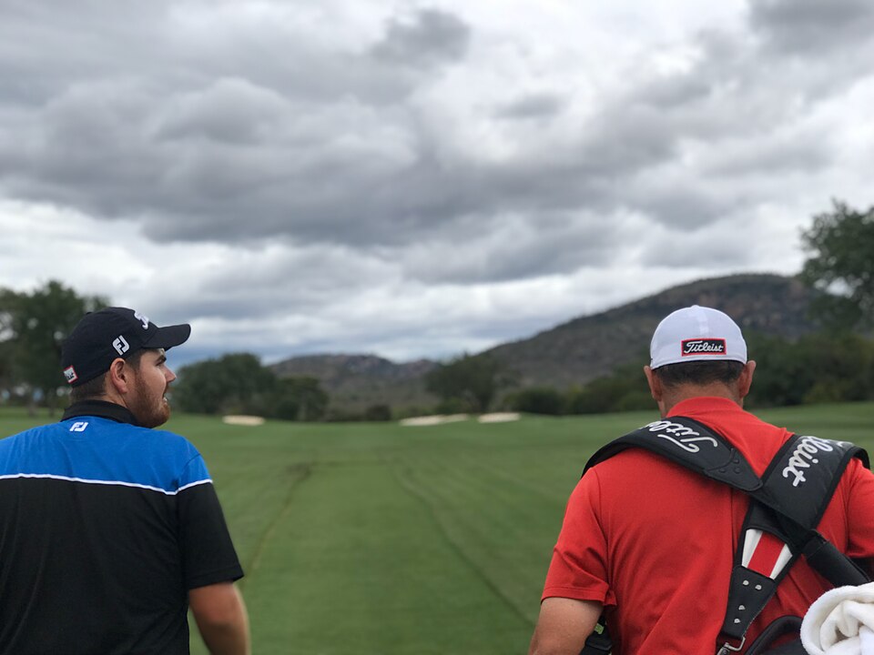 A professional golfer and his caddie, carrying the bag, walk away from the camera down a fairway under a cloudy sky, with bushveld hills ahead.