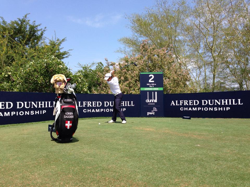 A professional golfer follows through on the 2nd tee at the Alfred Dunhill Championship, with tournament branding boards, a hole sign and his tour bag beside the tee.