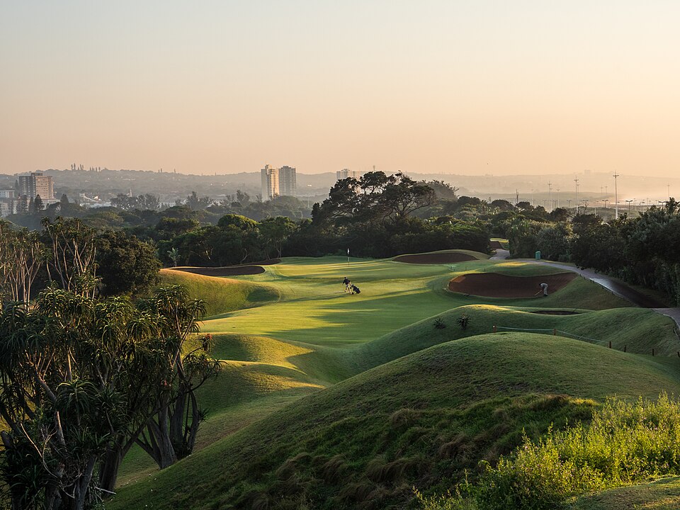 Dawn over a links-style course with steep grassy mounds and red-sand bunkers, where two greenkeepers are raking a bunker beside the green, with city buildings in the distance.