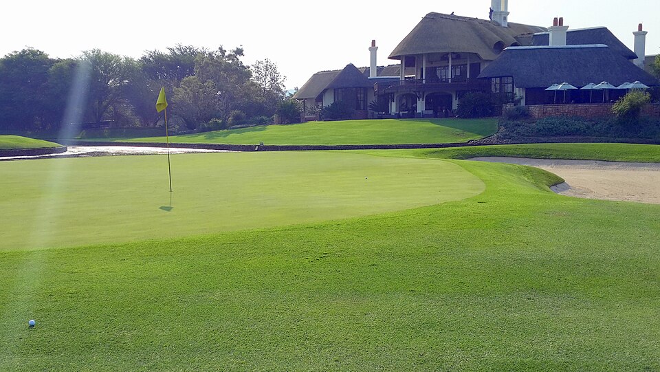 A thatched clubhouse with a terrace and umbrellas on a rise behind the 9th green, with a yellow flag on the green and water and a bunker in front.