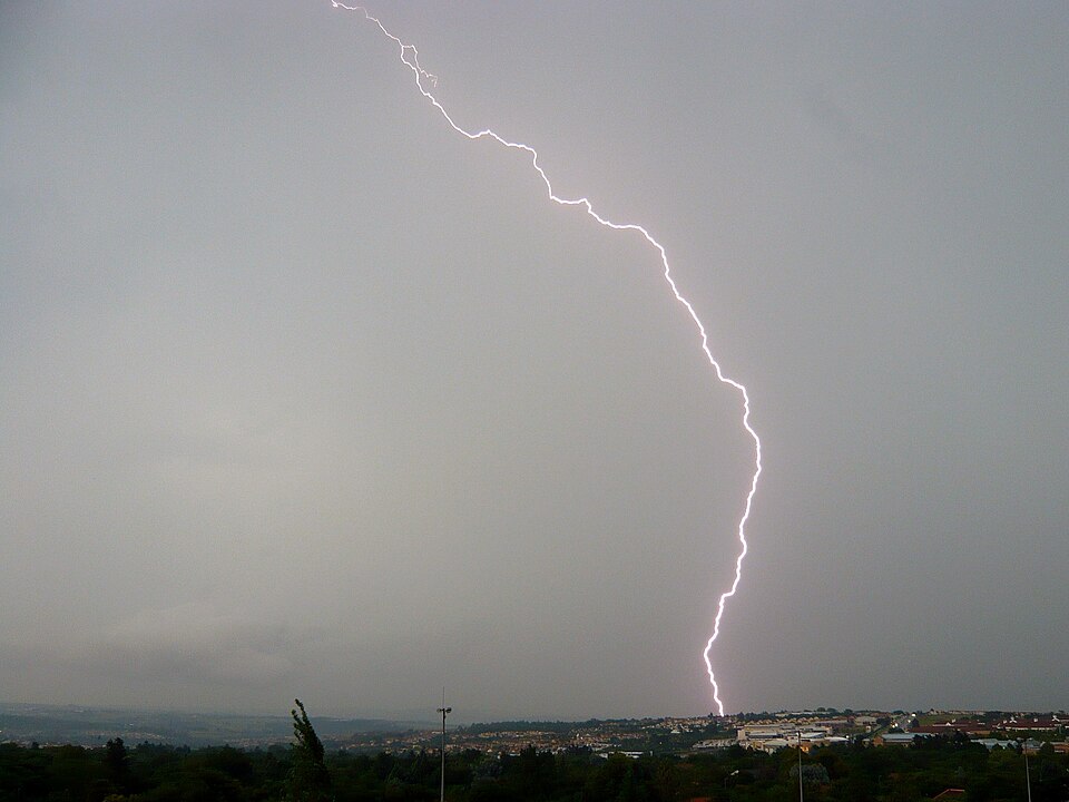 A single bright lightning bolt striking down from a grey sky onto the Johannesburg skyline at dusk.