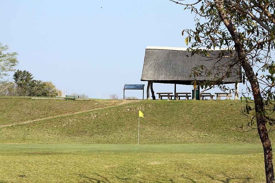 An unfenced green with a yellow flag in dry bushveld grass, below a thatched shelter on a rise, at Skukuza in the Kruger National Park.