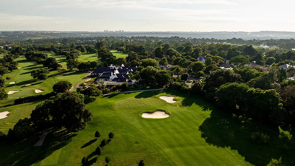 Aerial view over a tree-lined parkland golf course to a large clubhouse among the trees, with the Johannesburg skyline on the horizon.